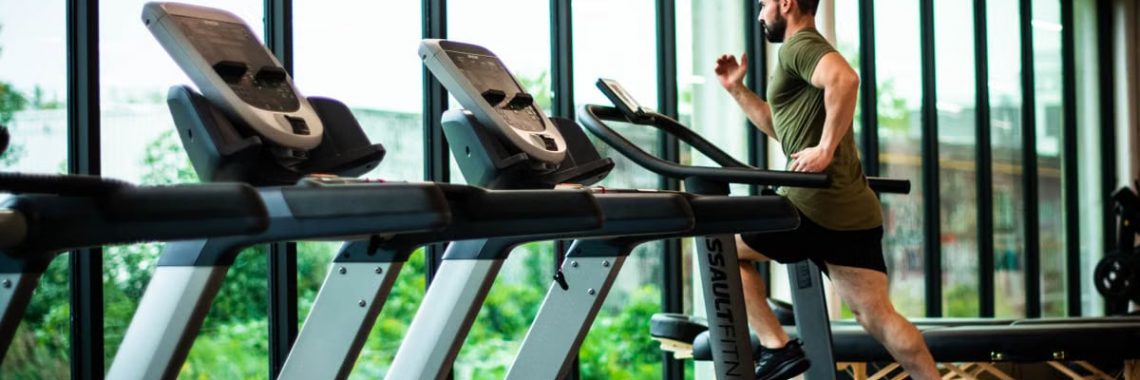 Young man workouts on treadmill in modern gym with large windows and natural light.